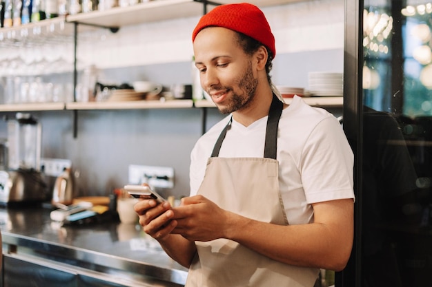 Un jeune barista portant un chapeau rouge sourit et utilise un smartphone au travail