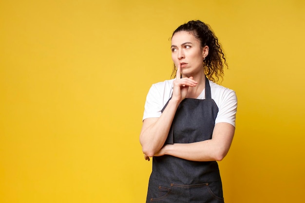 Une jeune barista réfléchie en uniforme se dresse sur un fond jaune et pense