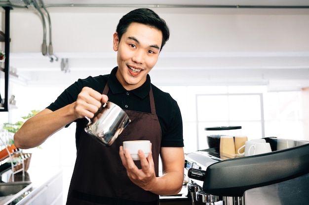 Jeune barista de sexe masculin portant un tablier, versant du lait chaud dans un expresso chaud.