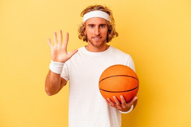 Jeune basketteur homme caucasien isolé sur fond jaune souriant joyeux montrant le numéro cinq avec les doigts.