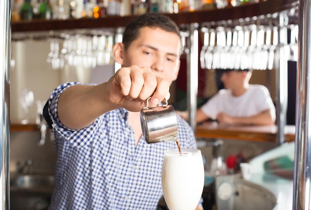 Jeune beau barman tient un jigger en acier inoxydable ou une tasse à mesurer et verse de la liqueur de café dans le verre de lait. L'accent est mis sur la main avec un jigger et un verre.