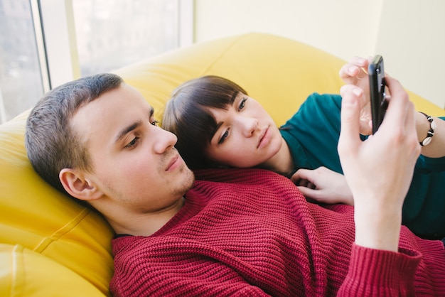 Jeune beau couple garçon et fille se reposant sur une chaise confortable et regarder des vidéos et des photos sur votre téléphone.