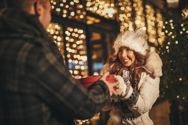 Jeune beau couple joyeux célébrant Noël dans la rue de la ville et se donnant des cadeaux.