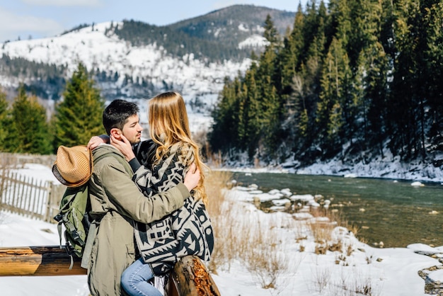 Jeune beau couple séjournant près de la rivière de montagne d'hiver, câlin et sourire. Espace libre pour le texte disponible