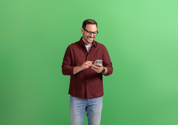 Un jeune et beau entrepreneur en lunettes travaillant à distance sur un téléphone intelligent sur un fond vert