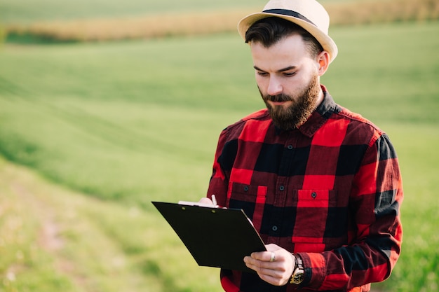 Jeune beau fermier barbu avec dossier debout dans un champ de blé vert au début de l&#39;été