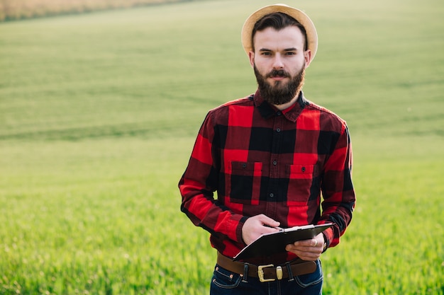 Jeune beau fermier barbu avec dossier debout dans un champ de blé vert au début de l&#39;été