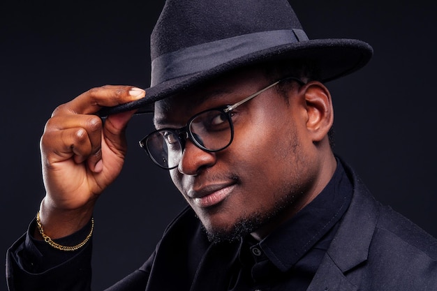 jeune beau garçon mélancolique afro-américain dans une veste et des lunettes sur fond noir dans le studio.mystère et chagrin se cachent