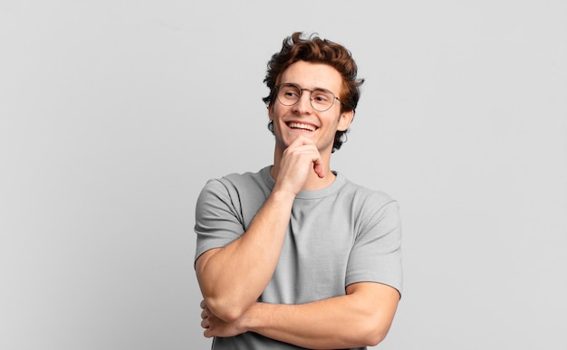 Jeune beau garçon souriant avec une expression heureuse et confiante avec la main sur le menton, se demandant et regardant sur le côté