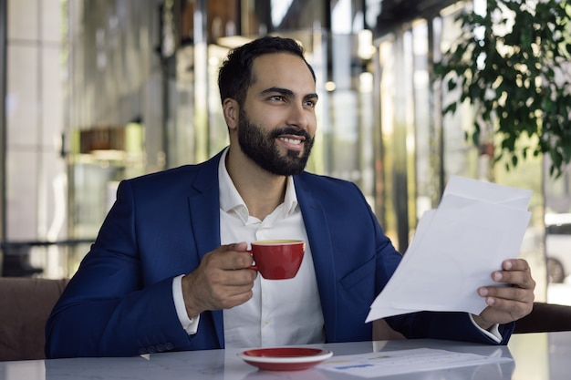 Jeune beau gestionnaire arabe travaillant avec des documents, assis sur le lieu de travail, tenant une tasse de café