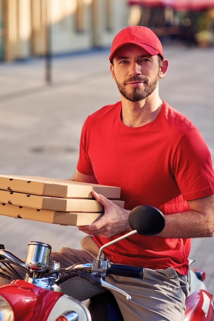 Jeune beau livreur caucasien en uniforme rouge sur scooter tenant des boîtes avec pizza et