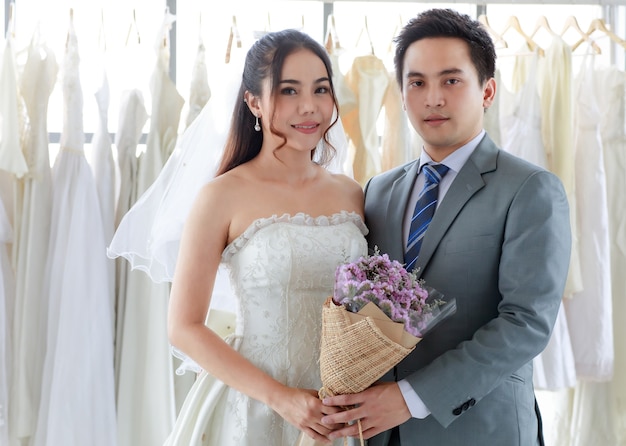 Jeune beau marié asiatique en costume formel gris avec cravate debout regarde la caméra avec une belle mariée heureuse en robe de mariée longue blanche tenant la main et un bouquet de fleurs ensemble dans le dressing.