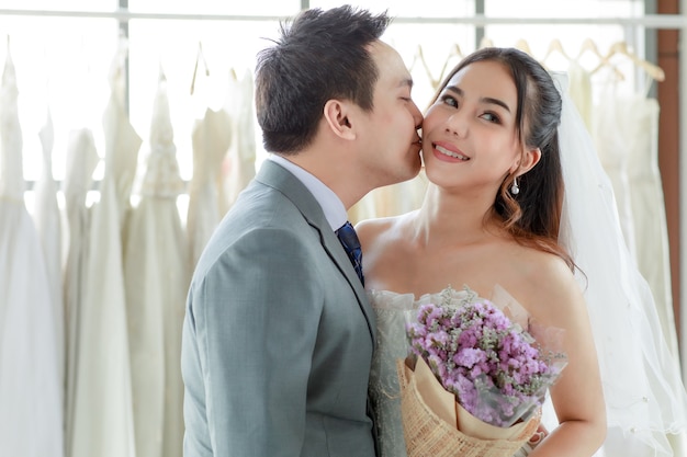 Jeune beau marié asiatique en costume gris avec cravate debout avec une belle mariée heureuse en robe de mariée longue blanche tenant la main et un bouquet de fleurs ensemble dans le vestiaire et embrasser sur la joue.