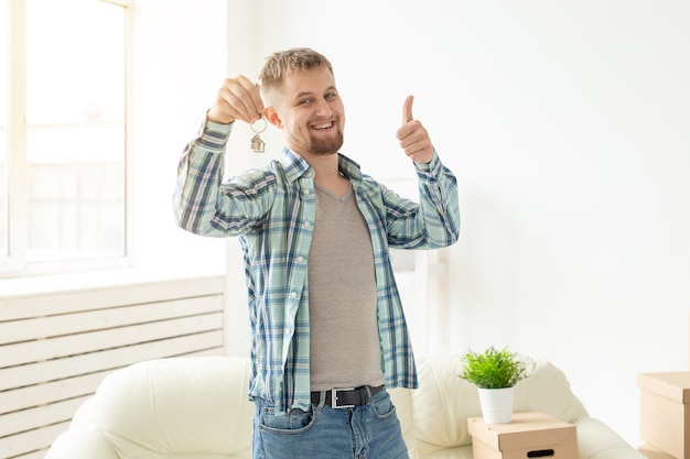 Jeune beau mec montre les clés de son nouvel appartement et montre les pouces vers le haut en se tenant debout dans le