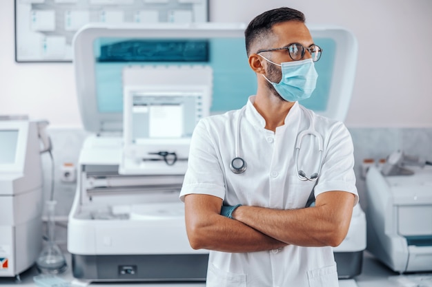 Jeune beau médecin debout à l'hôpital avec un masque facial et des gants en caoutchouc avec les bras croisés et en détournant les yeux.