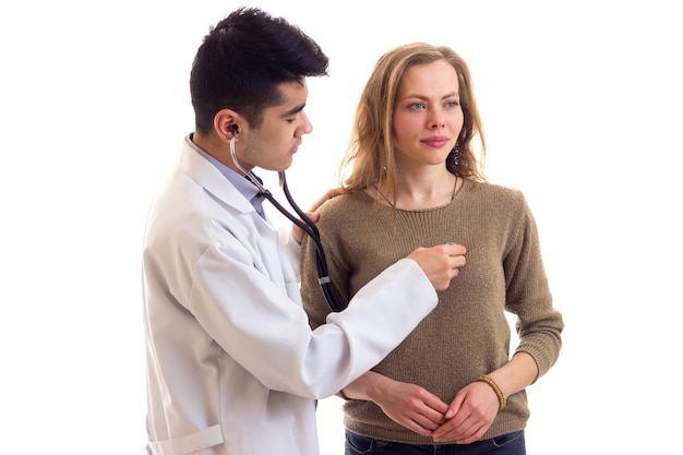 Jeune beau médecin en robe blanche avec stéthoscope écoutant le battement de coeur de la jeune femme