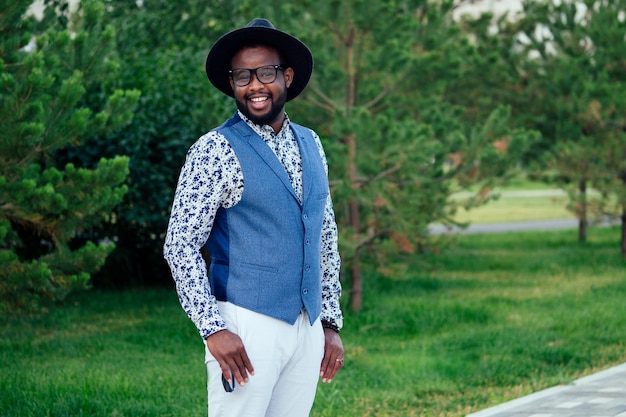 Un jeune et beau modèle afro-américain élégant dans un costume élégant, un pantalon blanc et un chapeau noir dans un parc d'été. homme d'affaires hispanique latino-américain mec noir posant au photoshoot.