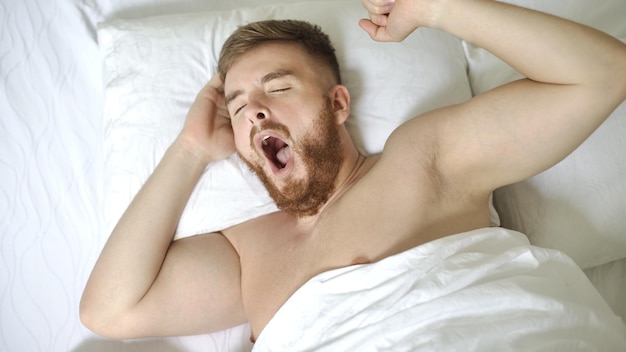 Photo jeune beau paresseux mec fatigué bâille dans son lit dans la chambre tôt le matin en étirant les mains du corps