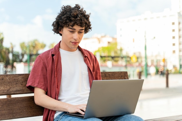 Jeune beau pigiste assis dans la rue pendant son temps libre avec un nouvel ordinateur portable moderne