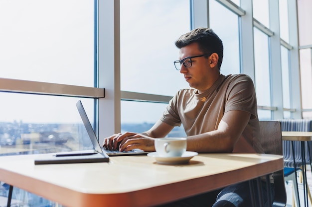 Jeune beau pigiste intelligent souriant à lunettes travaille sur un ordinateur portable tout en passant du temps dans un café Un étudiant positif est assis à une table en bois dans un café Heureux homme assis tout en travaillant à l'intérieur