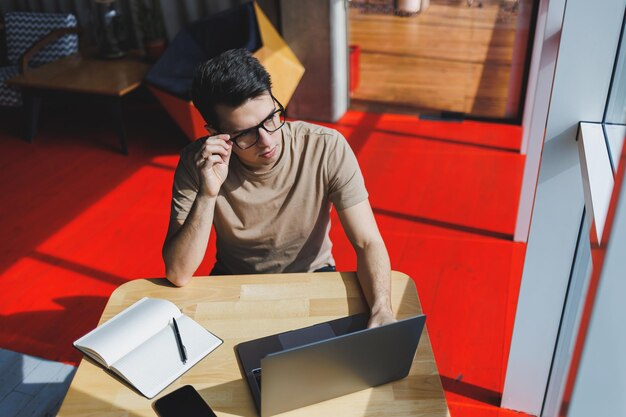 Jeune beau pigiste intelligent souriant à lunettes travaille sur un ordinateur portable tout en passant du temps dans un café Un étudiant positif est assis à une table en bois dans un café Heureux homme assis tout en travaillant à l'intérieur