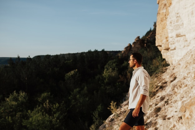 Jeune beau voyageur dans une chemise blanche et un short debout sur une pierre avec une belle vue sur la forêt de pins