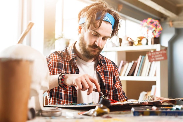 Jeune bel artiste contemporain masculin travaillant avec un couteau à palette et de l'huile dans son studio ensoleillé