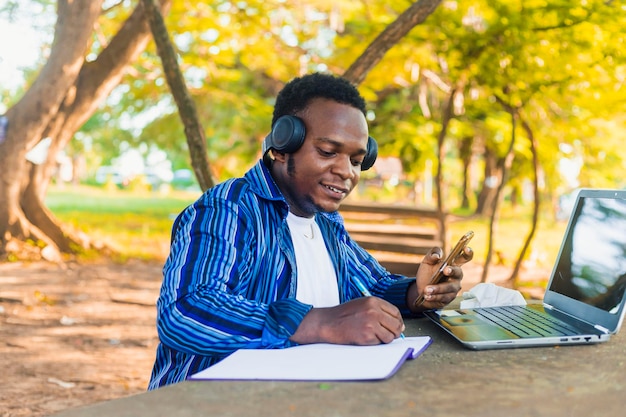 Jeune bel étudiant noir assis écrivant sur papier à l'aide d'un téléphone portable à la main