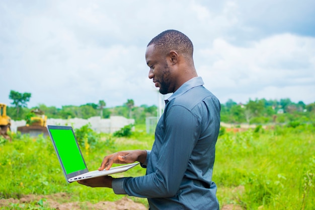 Jeune bel homme d'affaires africain noir debout sur une terre agricole à l'aide de son ordinateur portable.