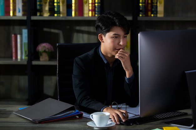 Jeune et bel homme d'affaires asiatique en costume assis dans une salle de travail privée et à la recherche d'un écran d'ordinateur