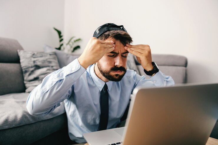 Photo jeune bel homme d'affaires barbu habillé décontracté assis à la maison et tenant la tête parce qu'il a mal à la tête. en face du bureau se trouve un ordinateur portable.