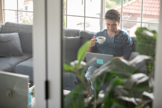 Jeune et bel homme d'affaires caucasien portant un tissu décontracté assis joyeusement sur un canapé, tenant une tasse et buvant du café et utilisant un ordinateur portable. Idée pour travailler à la maison, travail d'appel vidéo.