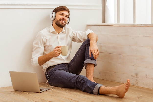 Jeune bel homme d'affaires en chemise classique blanche et jeans à l'aide d'un ordinateur portable écoutant de la musique tenant une tasse et souriant tout en étant assis sur le plancher en bois