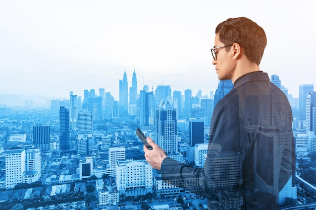 Jeune bel homme d'affaires en costume et lunettes utilisant le téléphone et pensant comment résoudre le problème de nouvelles opportunités de carrière MBA Kuala Lumpur en arrière-plan Double exposition