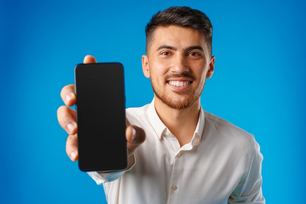 Jeune bel homme d'affaires montrant l'écran du smartphone noir sur fond bleu