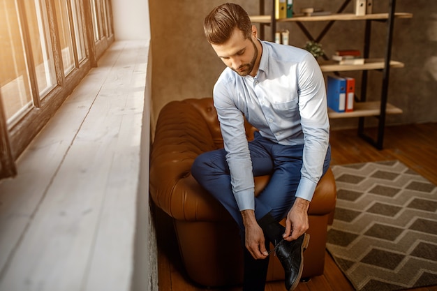 Jeune bel homme d'affaires s'asseoir sur le canapé et attacher les lacets sur les chaussures dans son propre bureau. Mec confiant à côté de la fenêtre. La lumière du soleil brille.