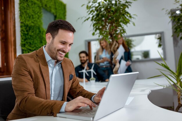 Jeune bel homme d'affaires souriant et prospère analysant des plans graphiques qui montrent le succès et travaillant sur un ordinateur portable au bureau.