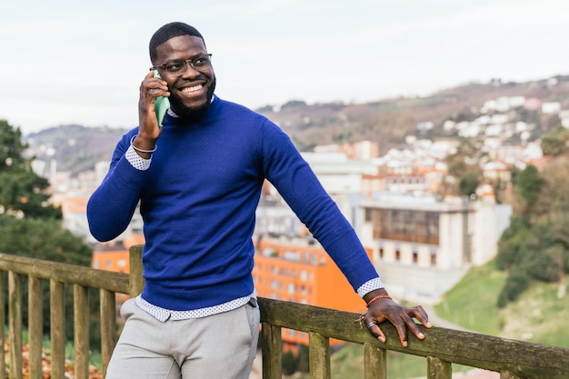 Jeune bel homme africain en pull bleu décontracté parlant au téléphone surplombant la ville depuis la balustrade du parc avec un éclairage époustouflant