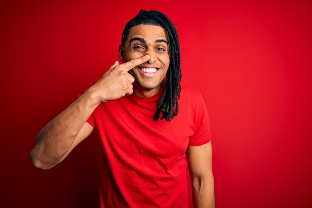 Jeune bel homme afro-américain afro avec des dreadlocks portant un t-shirt décontracté rouge pointant avec le doigt de la main vers le visage et le nez souriant concept de beauté joyeux