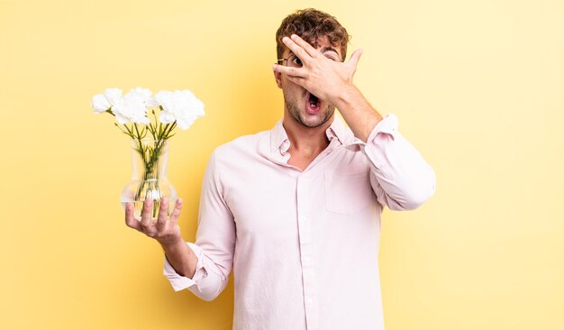 Jeune bel homme à l'air choqué, effrayé ou terrifié, couvrant le visage avec le concept de fleurs à la main