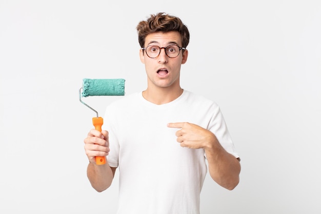 Jeune Bel Homme à L'air Choqué Et Surpris Avec La Bouche Grande Ouverte, Pointant Vers Lui-même Et Tenant Un Rouleau De Peinture