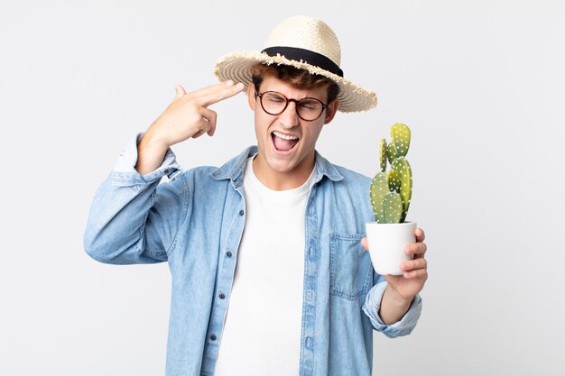 Jeune bel homme à l'air malheureux et stressé, geste de suicide faisant signe d'arme à feu. agriculteur tenant un cactus décoratif