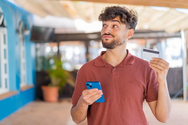 Jeune bel homme arabe à l'extérieur achetant avec le mobile avec une carte de crédit tout en pensant