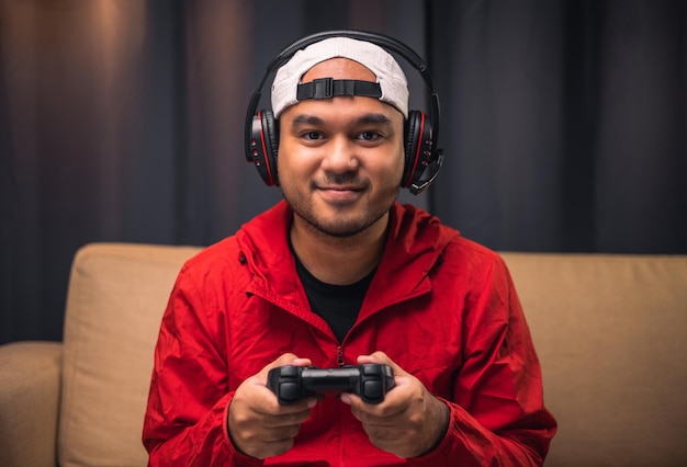 Jeune bel homme asiatique assis sur un canapé tenant un joystick dans le salon Happiness Streamer Homme indien portant un casque jouant à un jeu en ligne dans la chambre noire