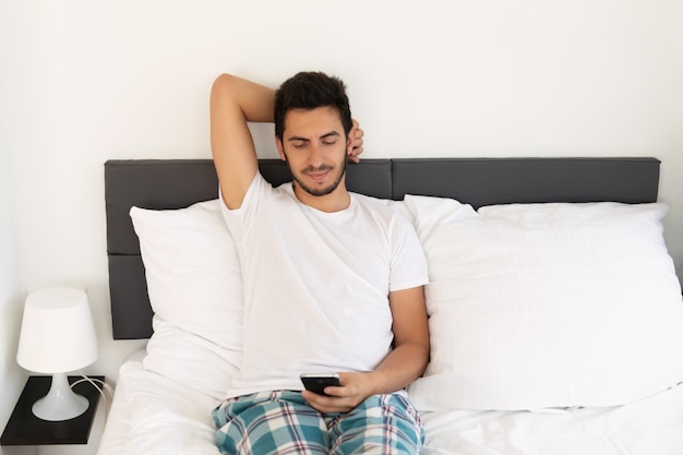 Jeune bel homme assis sur le lit. Il utilise un téléphone portable.