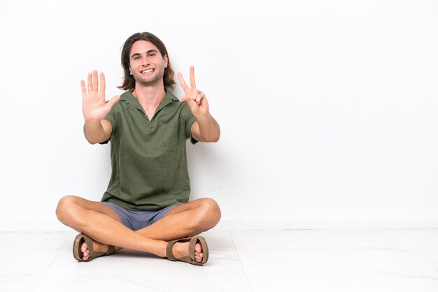 Jeune bel homme assis sur le sol isolé sur fond blanc comptant huit avec les doigts