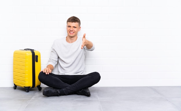 Jeune bel homme assis sur le sol avec une valise handshaking après bonne affaire