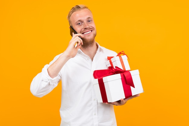 Jeune bel homme aux cheveux blonds en chemise blanche détient une boîte blanche avec cadeau et appeler le téléphone isolé sur jaune