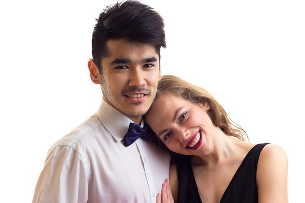 Jeune bel homme aux cheveux noirs en chemise blanche avec noeud papillon noir et belle jeune femme aux longs cheveux blonds en robe noire tenant un câlin rouge sur fond blanc en studio.