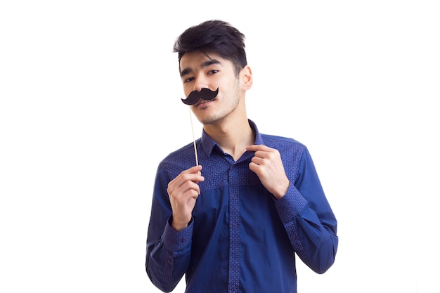Jeune bel homme aux cheveux noirs en chemise bleue tenant un bâton en carton de moustache en studio